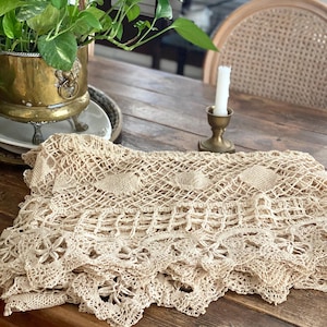 Può includere: Una tovaglia in pizzo all'uncinetto color crema, con bordo smerlato, appoggiata su un tavolo di legno. Il pizzo presenta motivi floreali e geometrici. Sono visibili anche un candeliere in ottone e una pianta in vaso.