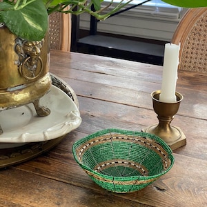May include: A small, round, beaded bowl with a green, gold, and brown color scheme. The bowl is made of small beads woven together to create a textured surface. The bowl is sitting on a wooden table.