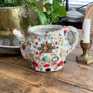 May include: A white ceramic teapot covered in a mosaic of colorful beads, buttons, and small metal charms. The surface features a textured, cracked finish with embedded red, blue, yellow, and green accents, a central brass flower piece, and a handle.