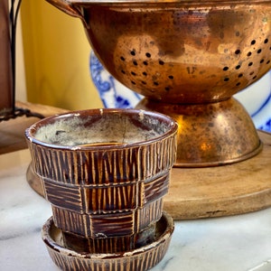 Könnte beinhalten: Ein kleiner, brauner Keramik-Blumentopf mit passender Untertasse. Der Topf hat ein strukturiertes Korbgeflecht-Design. Dahinter befindet sich ein Kupfer-Gemüsesieb, gefüllt mit Weinkorken, was auf eine Wein-Dekoration hindeutet.