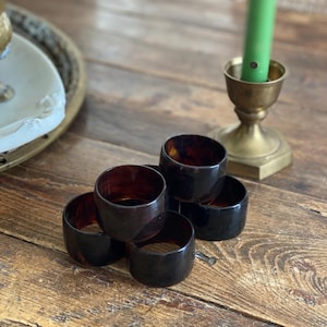 May include: A stack of six dark brown napkin rings with a tortoiseshell pattern. The rings are cylindrical and sit on a wooden surface. A brass candlestick with a green candle is in the background.