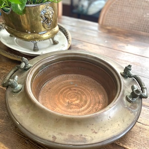 May include: Antique brass planter with decorative handles. The planter has a wide, shallow bowl shape with a circular design in the center. A second brass planter with a lion's head design is visible in the background.
