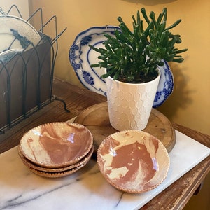 Puede incluir: Una colección de cuencos pequeños de cerámica hechos a mano con un diseño veteado en marrón rojizo y crema. Los cuencos tienen un borde texturizado y festoneado. Una pequeña planta suculenta en maceta se encuentra cerca, junto con platos antiguos azules y blancos.