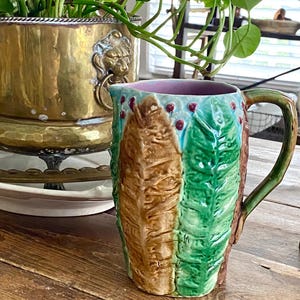 Puede incluir: Una jarra de cerámica vintage con un diseño de hojas marrones, verdes y azules. La jarra tiene un asa y un interior morado. La jarra está sobre una mesa de madera.