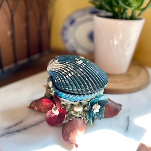 Peut inclure: Boîte décorative en forme de coquillage avec couvercle à charnière. La boîte est turquoise et blanche, avec une surface texturée. La base est ornée d'éléments décoratifs rouges et bleus, et de petits coquillages blancs.