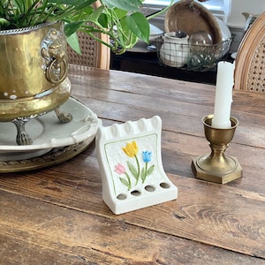 May include: A white ceramic toothbrush holder with a floral design of yellow, blue, and pink tulips. The holder has four holes for toothbrushes and a scalloped top. A brass candle holder with a white candle is also visible.