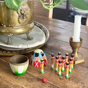 May include: A still-life arrangement on a wooden table features a brass planter with a lion's head, a candle holder, and a vintage toy set. The toy set includes miniature toy soldiers, a red ball, and a painted egg-shaped container.