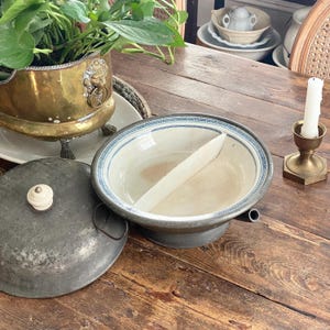 Könnte beinhalten: Eine geteilte Vintage-Servierschale mit blau-weißem Muster am Rand, auf einem Holztisch. Ein Metalldeckel mit weißem Knopf liegt in der Nähe. Ein Messingtopf mit Grün und ein Kerzenhalter sind ebenfalls sichtbar.