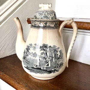 Puede incluir: Una tetera vintage de color crema con un diseño floral en blanco y negro. La tetera tiene una tapa con un pomo en la parte superior y un pico en el lateral. La tetera está sobre una superficie de madera.