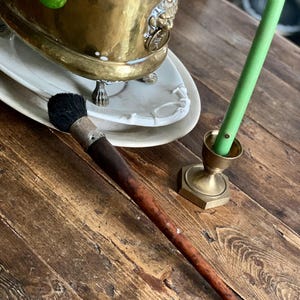 Może przedstawiać: Martwa natura w stylu vintage z pędzlem z długim trzonkiem i czarną główką z włosia, zieloną świecą w mosiężnym świeczniku i mosiężną donicą z rośliną. Obiekty spoczywają na zwietrzałej drewnianej powierzchni.