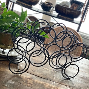 May include: A black metal wine rack with a circular design, holding multiple bottles. The rack sits on a rustic wooden table, with a plant and shelves in the background. The rack is designed to hold several bottles of wine.