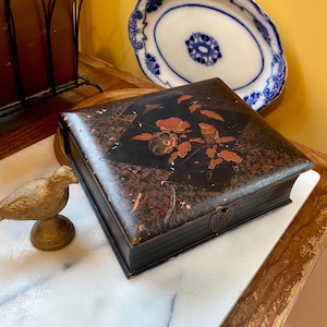 May include: A black and brown antique box with a floral design and a clasp. A gold-colored bird figurine sits nearby. A white and blue oval plate is in the background.