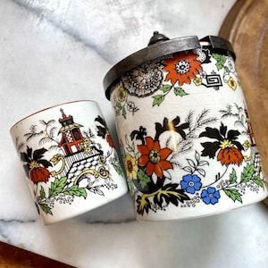 Könnte beinhalten: Zwei antike Keramikbehälter mit floralen und architektonischen Designs. Der größere Behälter hat einen Metallrand und einen Deckel. Die Behälter weisen orange, schwarze, blaue und grüne Blumenmuster auf weißem Hintergrund auf.