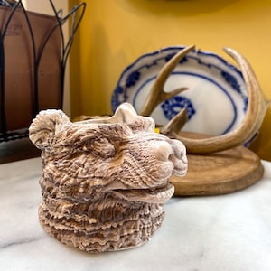 Puede incluir: Una maceta con forma de oso de color beige, con detalles tallados en la cara y el pelaje del oso. La maceta tiene una superficie texturizada y está colocada sobre una superficie de mármol blanco.