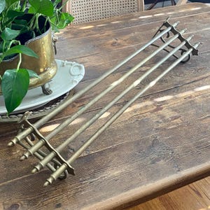 May include: A long, horizontal brass towel rack featuring three parallel rods connected by two decorative end brackets. Each bracket has four vertical finial-topped posts and a curved mounting hook, showcasing a metallic gold-toned finish.