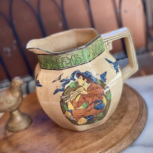 Puede incluir: Jarra de cerámica color crema con asa y boquilla. La jarra presenta un diseño pintado de una persona tocando un instrumento de viento, rodeada de pájaros azules y follaje verde. El borde tiene un borde verde con texto.