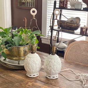 Puede incluir: Dos lámparas de mesa de cerámica blanca con un diseño floral. Las lámparas tienen cuello y base color latón y un remate blanco circular. Las lámparas están sobre una mesa de madera.