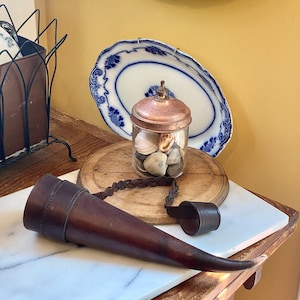 May include: A dark brown leather hunting horn with a braided strap and a flared bell. A glass jar with a copper lid filled with seashells sits on a wooden board. Decorative blue and white plates are in the background.
