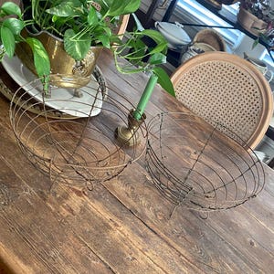 May include: Two antique wire baskets with a distressed finish sit on a weathered wooden table. A brass planter with green plants and a green candle holder are also on the table. The scene evokes a vintage, rustic aesthetic.