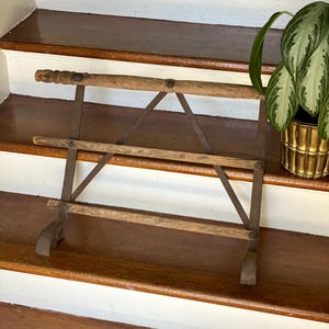 May include: An antique wooden and metal drying rack. The rack has three horizontal wooden bars supported by metal brackets. The top bar has a decorative, carved handle. The rack is displayed on wooden stairs.