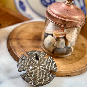 May include: A bronze sand dollar sits on a white surface. A glass jar with a copper lid filled with seashells and pebbles is behind it. A wooden chopping board and a blue and white plate are in the background.