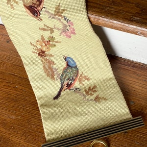 May include: Needlepoint wall hanging depicting two birds on flowering branches. The birds are brown and blue with detailed stitching. The background is a light yellow-green. The hanging has a wooden rod at the bottom and a gold ring.