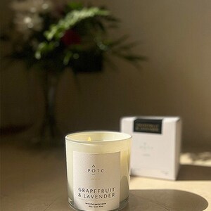 Op de afbeelding: Een witte kaars in een matglazen pot met een wit etiket waarop staat "A POTC Grapefruit & Lavendel" handgemaakte geurkaars 300g 12oz 80hrs.