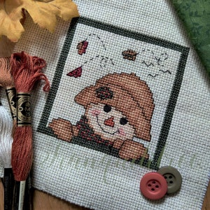 May include: A cross-stitch pattern of a smiling scarecrow with a brown hat and red scarf, surrounded by falling leaves. The pattern is framed in green and is on a white background. There are three skeins of embroidery floss and two buttons in the image.