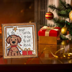 May include: A framed cross-stitch featuring a brown dog with Christmas lights and a red gift box. The text reads "Dear Santa, I tried to be good." A red and gold gift box and Christmas tree ornaments are in the background.
