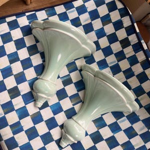 May include: Two matching pale mint green ceramic wall sconces featuring a flared, trumpet-shaped top with scalloped edges and a tapered, bulbous base. The smooth, glossy finish highlights the curved, architectural silhouette of these wall-mounted pieces.