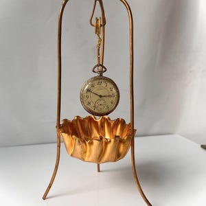 May include: A gold-toned metal stand with a curved top and three legs, featuring a suspended round pocket watch with a white face, black numerals, and a small sub-dial. Below the watch sits a gold-toned, ruffled, bowl-shaped metal dish.