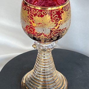 May include: A ruby red wine glass with gold leaf and grape vine designs. The rim and base are trimmed with gold. The stem is clear with gold accents. The glass is approximately 6 inches tall.