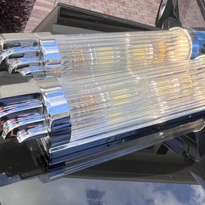 May include: Two vintage, clear glass and chrome light fixtures. Each fixture has a ribbed glass tube with chrome end caps. The fixtures are resting on a dark, reflective surface.