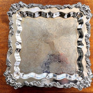 May include: A silver-colored, square serving tray with ornate, floral-style detailing. The tray has a decorative, wavy border and a detailed, embossed pattern across the surface. The edges feature elaborate scrollwork and floral designs, adding to its elegant appearance.