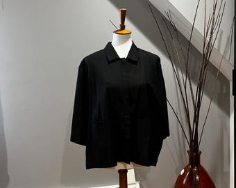 May include: A black, short-sleeved jacket with two front pockets displayed on a mannequin. The jacket has a collared neckline and a button-up front. The mannequin is on a wooden stand. A decorative vase with branches is to the right.