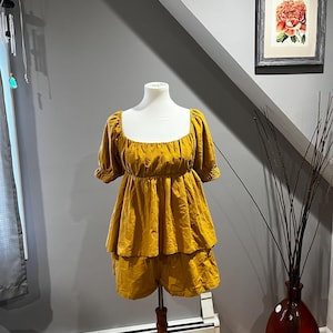 K&ouml;nnte beinhalten: Senfgelber Overall mit quadratischem Ausschnitt und kurzen &Auml;rmeln. Das Oberteil hat einen gerafften Oberteil und die Shorts sind gestuft. Das Outfit wird auf einer wei&szlig;en Schaufensterpuppe pr&auml;sentiert. Der Overall ist aus einem weichen, flie&szlig;enden Stoff.