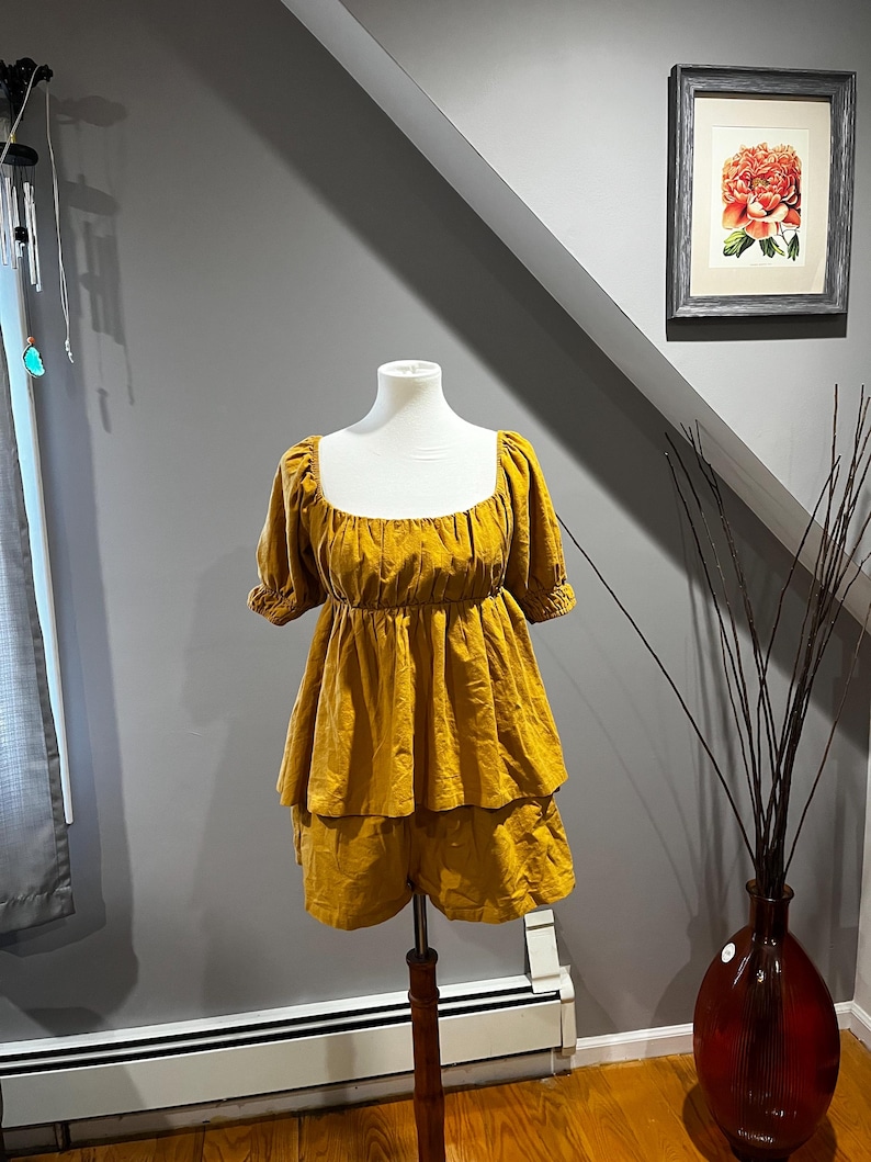 K&ouml;nnte beinhalten: Senfgelber Overall mit quadratischem Ausschnitt und kurzen &Auml;rmeln. Das Oberteil hat einen gerafften Oberteil und die Shorts sind gestuft. Das Outfit wird auf einer wei&szlig;en Schaufensterpuppe pr&auml;sentiert. Der Overall ist aus einem weichen, flie&szlig;enden Stoff.