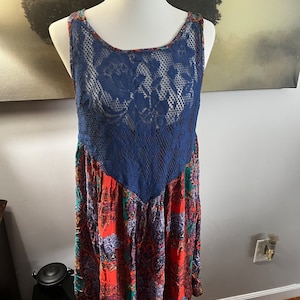 Puede incluir: Vestido sin mangas sobre un maniqu&iacute;. El vestido presenta un corpi&ntilde;o de encaje azul marino y una falda estampada con tonos rojos, verdes y morados. El vestido tiene un escote redondo y un estilo bohemio fluido.