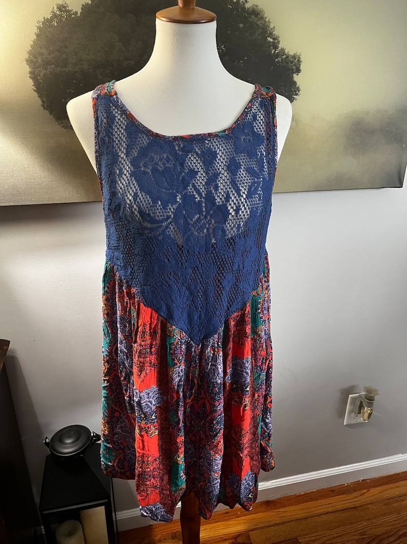 Puede incluir: Vestido sin mangas sobre un maniqu&iacute;. El vestido presenta un corpi&ntilde;o de encaje azul marino y una falda estampada con tonos rojos, verdes y morados. El vestido tiene un escote redondo y un estilo bohemio fluido.