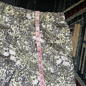 May include: White shorts with a green and gold floral print. The shorts have a waistband with a seam down the center back. The shorts are measured with a tape measure, showing a length of 17 inches.