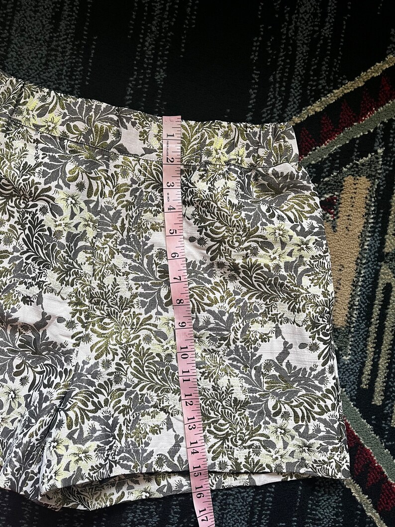 May include: White shorts with a green and gold floral print. The shorts have a waistband with a seam down the center back. The shorts are measured with a tape measure, showing a length of 17 inches.
