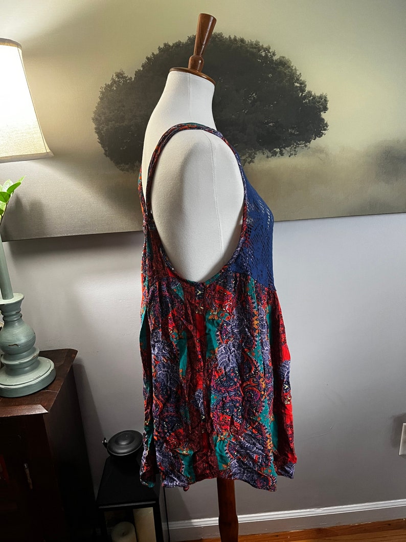 Puede incluir: Una blusa sin mangas con un vibrante estampado de cachemira en rojo, verde azulado y morado. La blusa presenta un canes&uacute; y tirantes de ganchillo azul marino. La prenda se exhibe en un maniqu&iacute;.