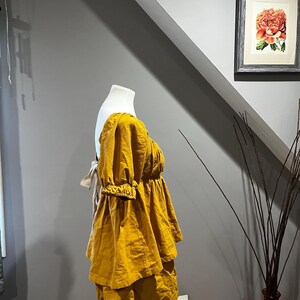 K&ouml;nnte beinhalten: Senfgelbes Leinen-Top und -Shorts-Set auf einer Schaufensterpuppe. Das Top hat einen gerafften Ausschnitt und kurze, gepuffte &Auml;rmel. Die Shorts haben ein gestuftes Design. Das Outfit steht vor einer grauen Wand mit einem gerahmten Blumendruck.