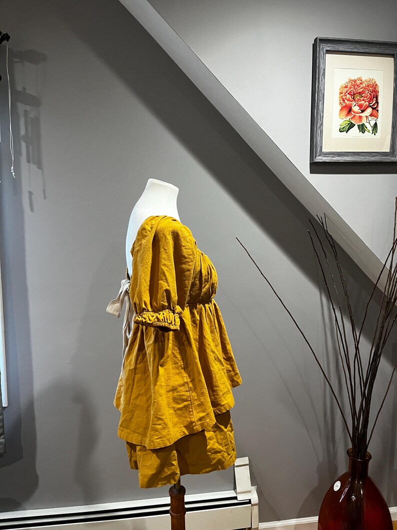K&ouml;nnte beinhalten: Senfgelbes Leinen-Top und -Shorts-Set auf einer Schaufensterpuppe. Das Top hat einen gerafften Ausschnitt und kurze, gepuffte &Auml;rmel. Die Shorts haben ein gestuftes Design. Das Outfit steht vor einer grauen Wand mit einem gerahmten Blumendruck.
