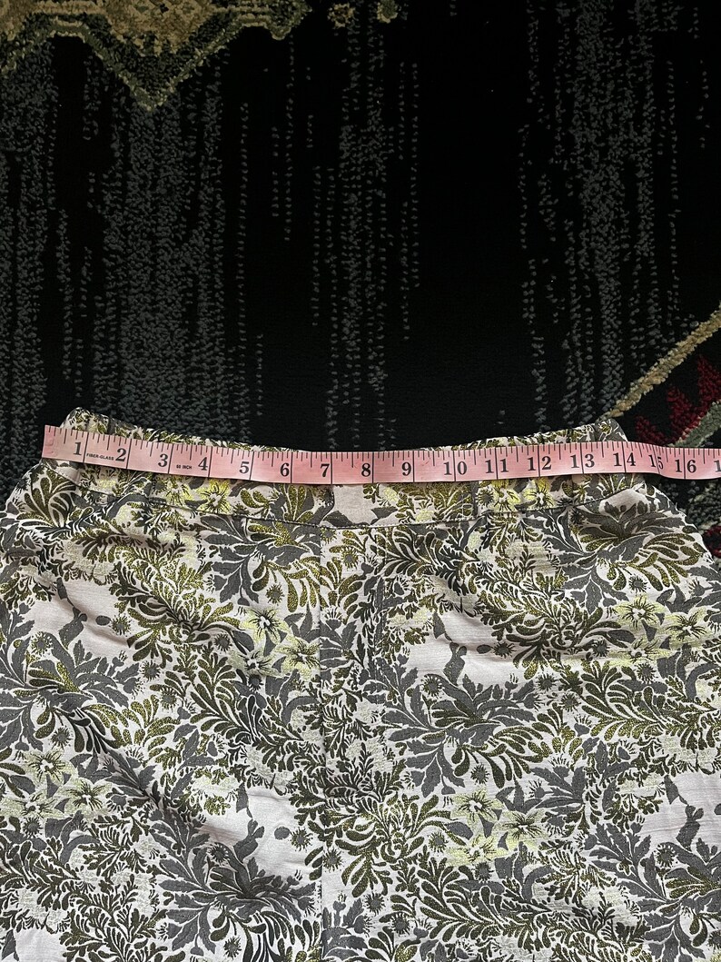 May include: A white fabric skirt with a green and gold floral pattern. The skirt has a waistband with a pink measuring tape that reads "1 2 3 4 5 6 7 8 9 10 11 12 13 14 15 16".