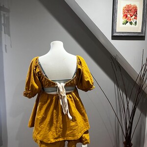 K&ouml;nnte beinhalten: Senfgelbes zweiteiliges Outfit auf einer Schaufensterpuppe. Das Oberteil hat kurze, gepuffte &Auml;rmel und einen tiefen R&uuml;cken mit cremefarbenen Bindeb&auml;ndern. Die Shorts sind geschichtet. Das Outfit wird auf einem Holzst&auml;nder pr&auml;sentiert. Ein gerahmter Blumendruck h&auml;ngt an der grauen Wand.
