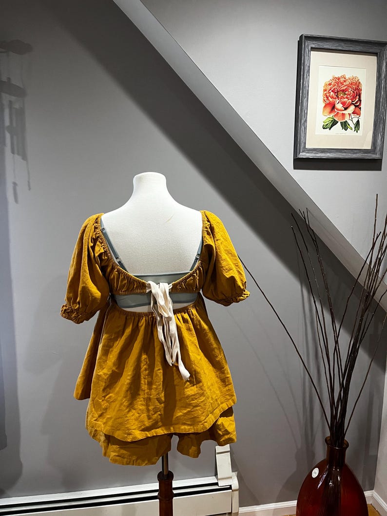 K&ouml;nnte beinhalten: Senfgelbes zweiteiliges Outfit auf einer Schaufensterpuppe. Das Oberteil hat kurze, gepuffte &Auml;rmel und einen tiefen R&uuml;cken mit cremefarbenen Bindeb&auml;ndern. Die Shorts sind geschichtet. Das Outfit wird auf einem Holzst&auml;nder pr&auml;sentiert. Ein gerahmter Blumendruck h&auml;ngt an der grauen Wand.