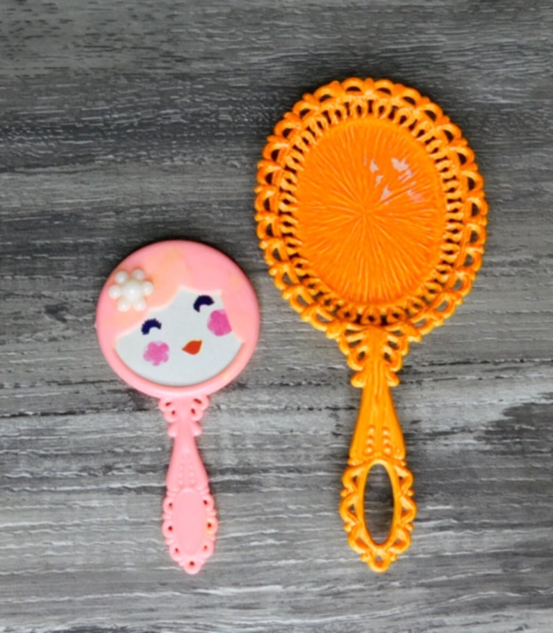 May include: Two hand mirrors, one orange with a decorative design and one pink with a cartoon face.
