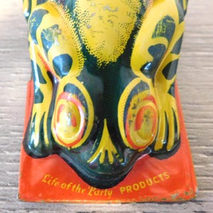 May include: A vintage tin frog figurine featuring a yellow and dark green mottled pattern with orange-rimmed eyes. The frog is positioned on a rectangular orange base with the text Life of the Party PRODUCTS printed in yellow on the front edge.