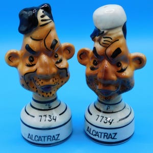 May include: A pair of ceramic salt and pepper shakers, each shaped like a cartoonish face with a hat. The faces are tan with black accents and a white base with the word "ALCATRAZ" and the number "7734".